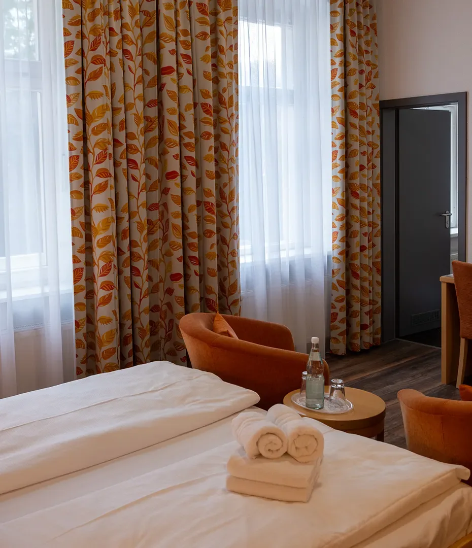 Hotelzimmer mit zwei Einzelbetten, orangen Sesseln, rundem Beistelltisch mit Wasserflasche und Gläsern sowie gemusterten Vorhängen
