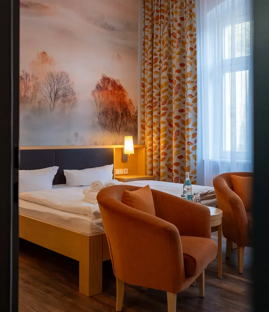 Hotelzimmer mit Doppelbett, zwei orangeroten Sesseln, kleinem runden Tisch und großer Fensterfront mit gemusterten Vorhängen