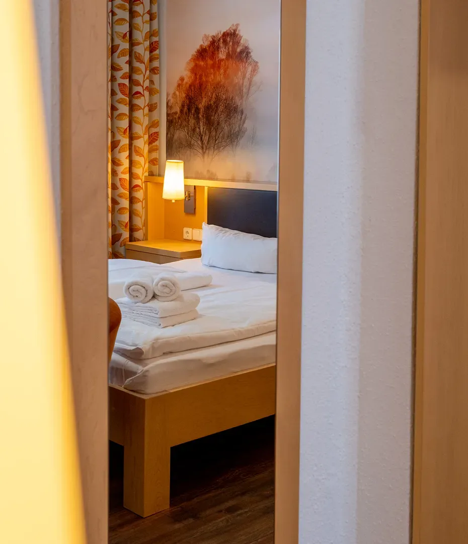 Blick durch eine Tür auf ein Doppelbett mit weißen Handtüchern und einem Bild mit Baum im Hintergrund
