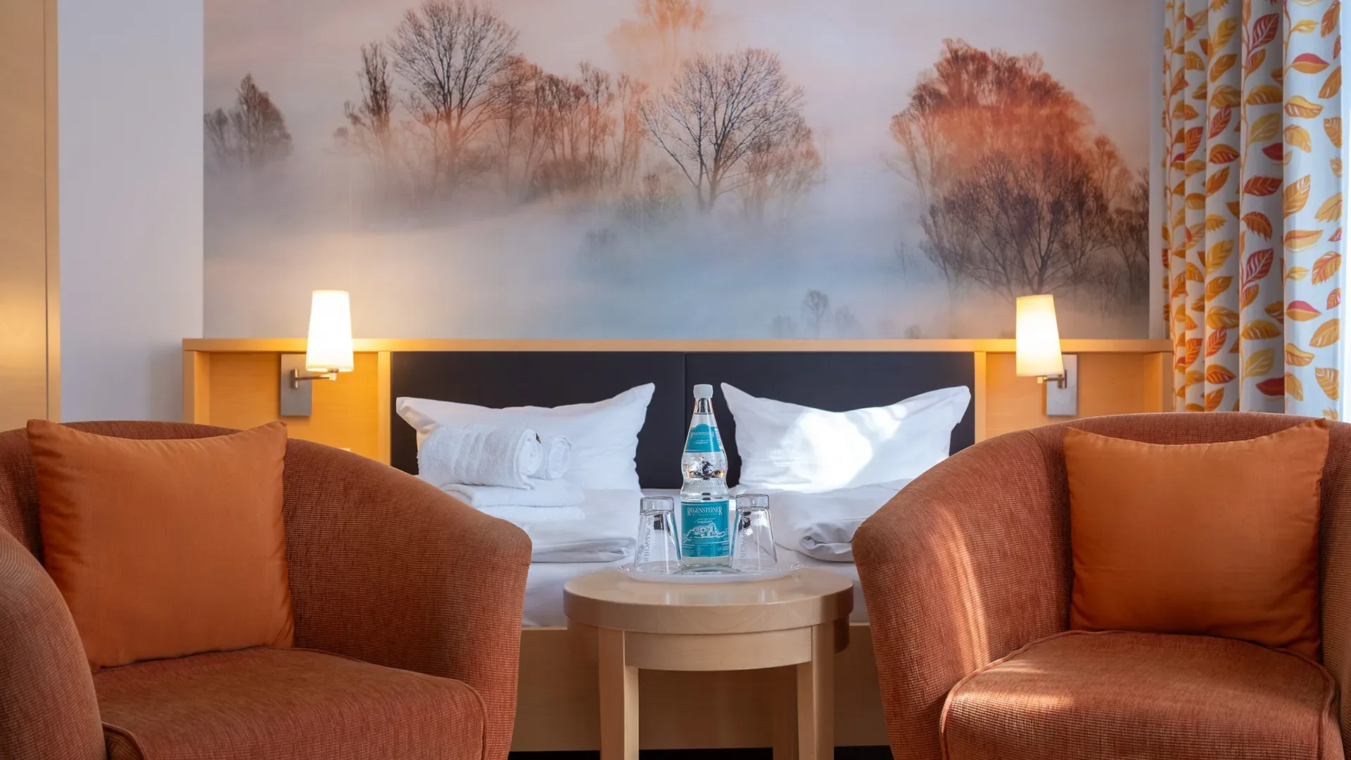 Hotelzimmer mit Doppelbett, zwei orangefarbenen Sesseln, Nachttisch mit Wasserflasche und Gläsern sowie Wandbild mit Bäumen im Nebel
