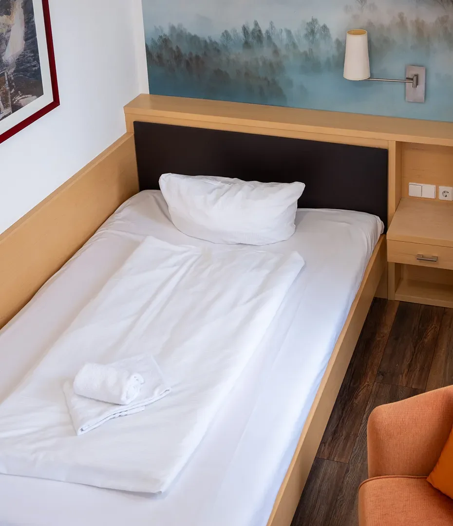 Einzelbett mit weißer Bettwäsche, einem Kissen und einem zusammengerollten Handtuch in einem Hotelzimmer mit Holzmöbeln und einem orangen Stuhl