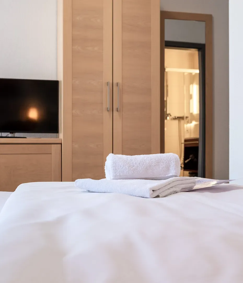 Einzelzimmer mit weiß bezogenem Bett, darauf liegen zwei gefaltete weiße Handtücher, im Hintergrund ein Holzschrank und ein Fernseher