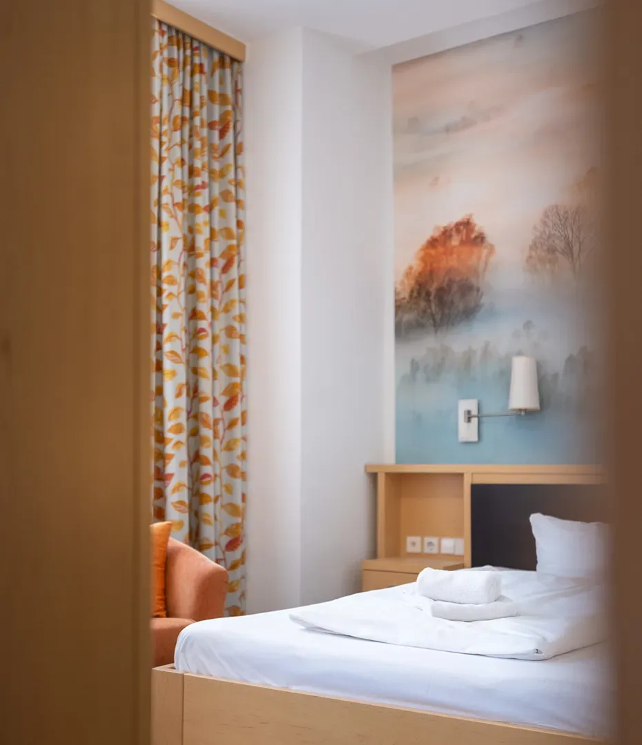 Einzelzimmer mit weiß bezogenem Bett, Handtüchern, einem Sessel und einer Wand mit Naturmotivtapete