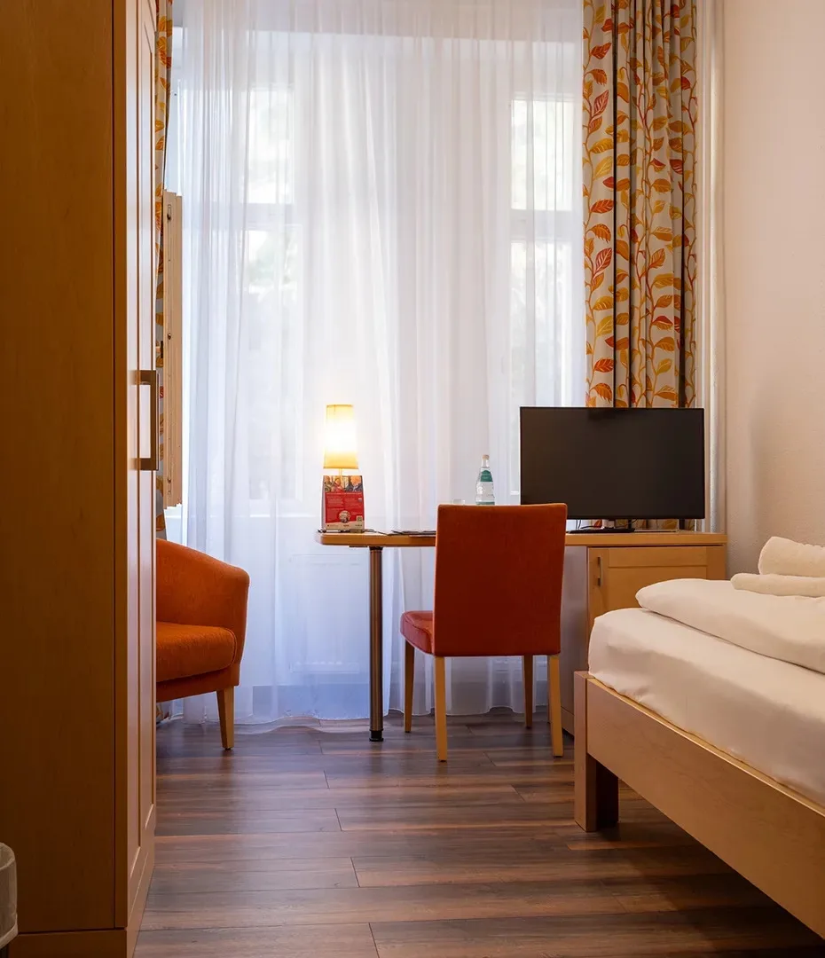 Einzelzimmer mit Bett, Schreibtisch, Stuhl, Fernseher und Fenster mit hellen Vorhängen