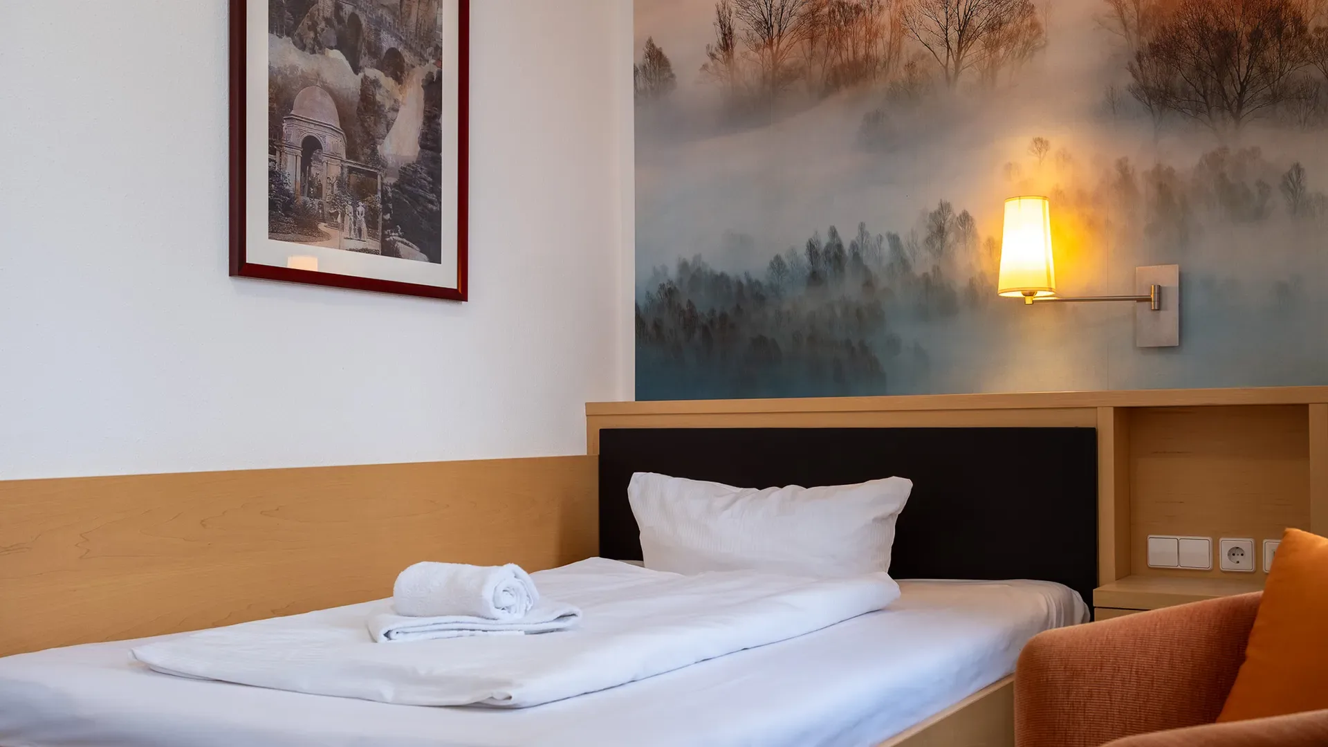 Einzelzimmer mit einem Bett, weißer Bettwäsche, einem Handtuch auf dem Bett, einer Wandlampe und einem Bild an der Wand