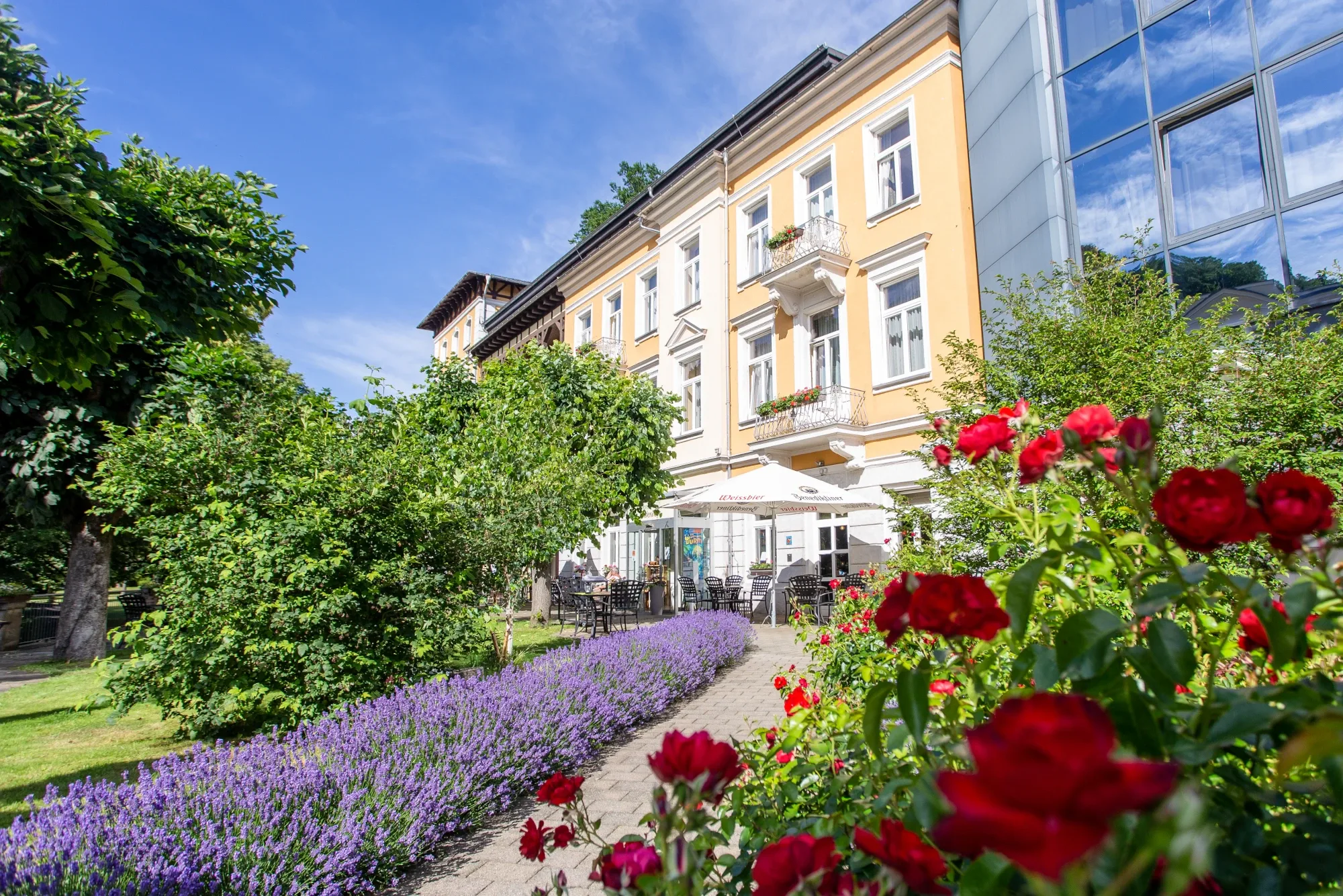 Außenansicht eines mehrstöckigen Hotels mit gelber Fassade, davor blühende Beete mit roten Rosen und lila Blumen sowie ein Sitzbereich mit Sonnenschirmen