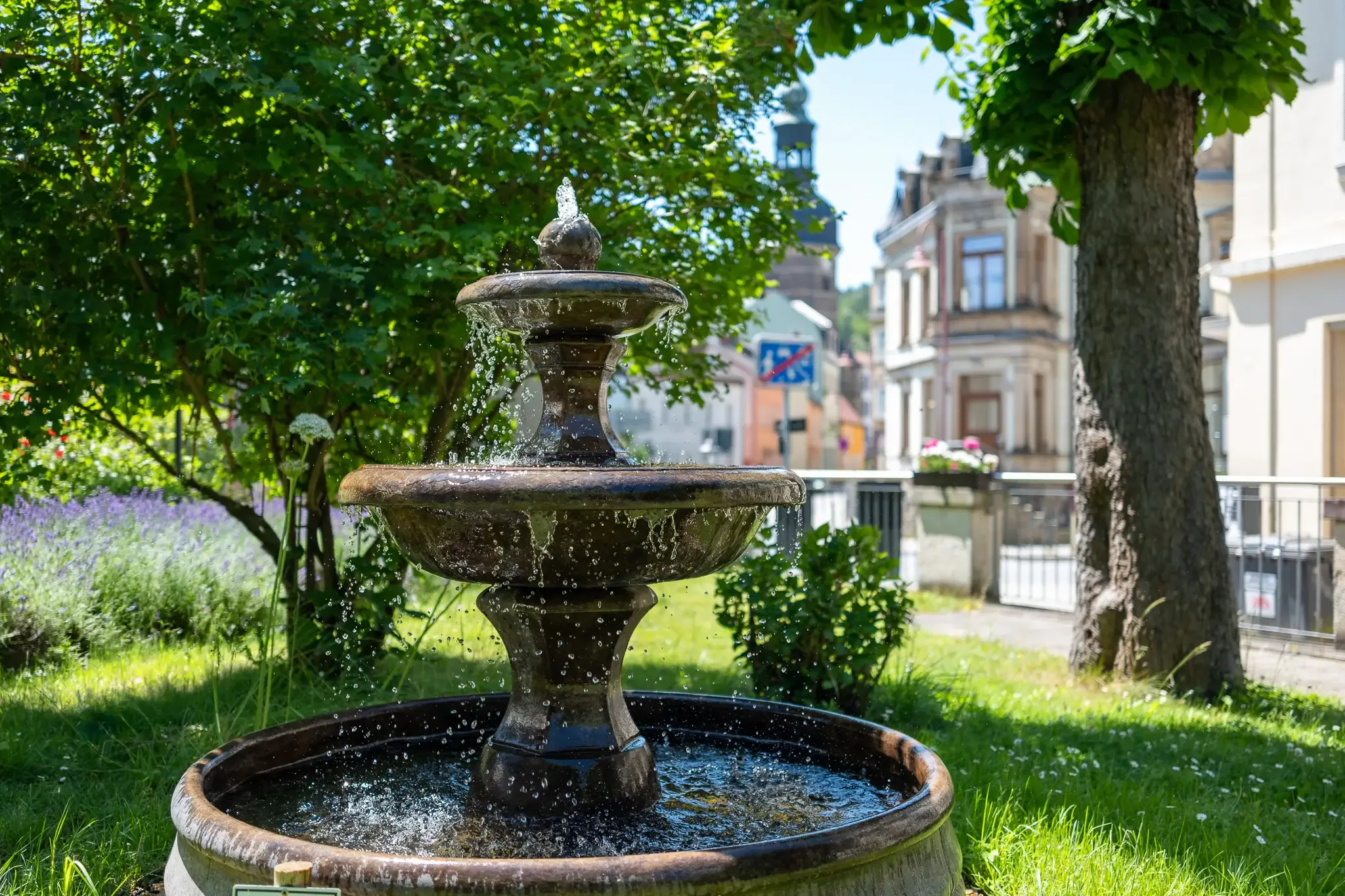 Zweistufiger Brunnen mit fließendem Wasser im Garten vor mehreren Gebäuden und Bäumen
