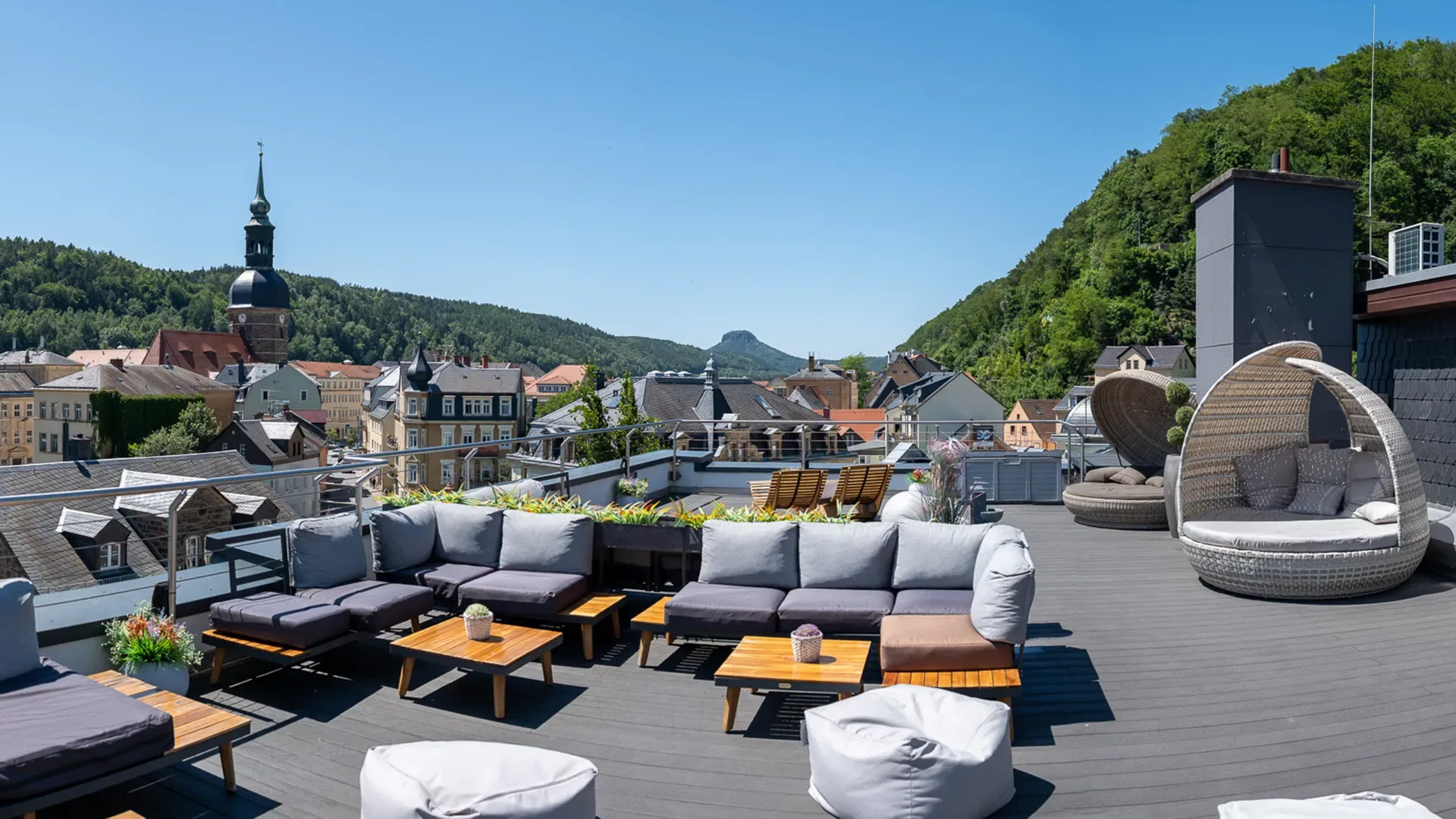 Dachterrasse mit Sitzgruppen aus Sofas, Sesseln und Tischen vor Stadt- und Bergkulisse