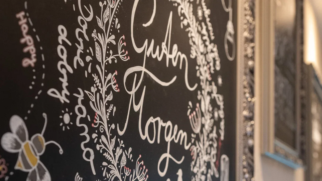 Handgemalte Kreidetafel mit „Guten Morgen"-Schriftzug, Blumen und Biene beim Frühstück im Lindenhof