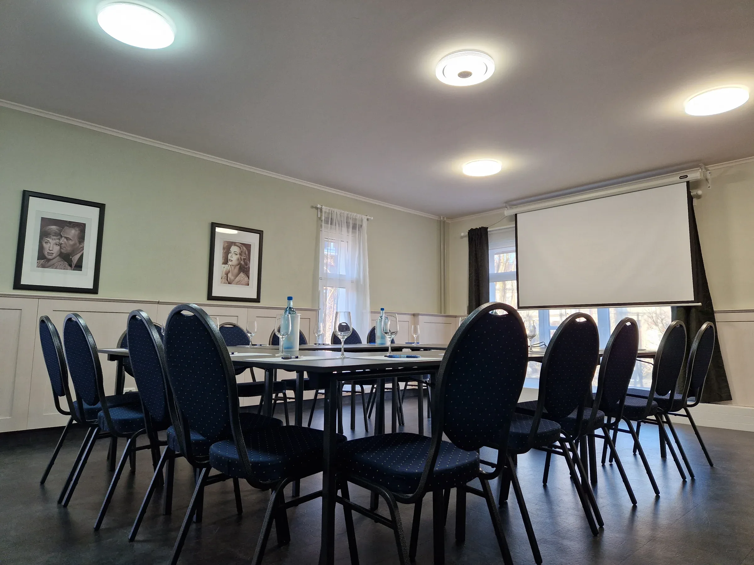 Lindenstube mit U-förmiger Tischaufstellung, blauen Polsterstühlen und heruntergelassener Leinwand für eine Tagung