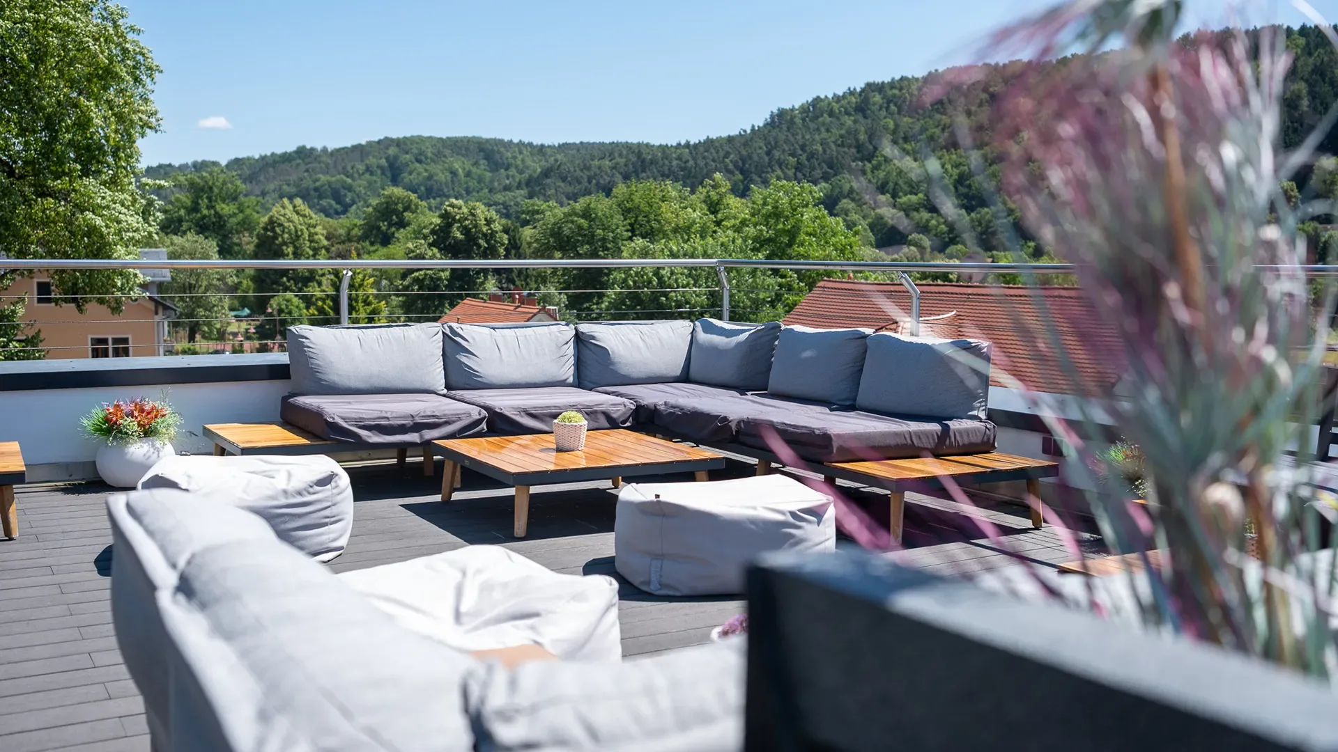 Terrasse mit grauer Polsterecke, Holztisch und Sitzsäcken vor bewaldeten Hügeln