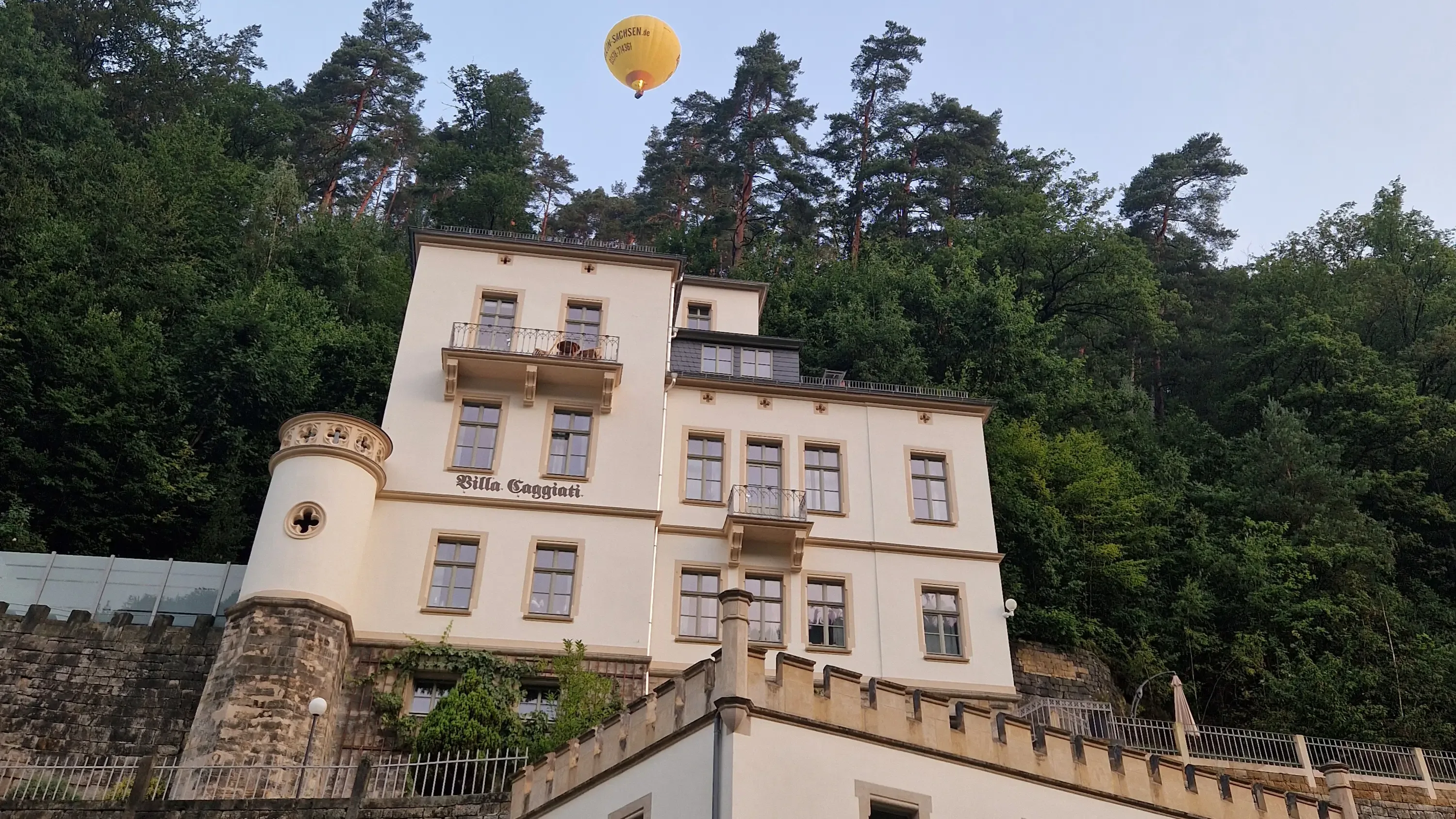 Mehrstöckiges historisches Gebäude mit Balkonen und Rundturm vor bewaldetem Hang