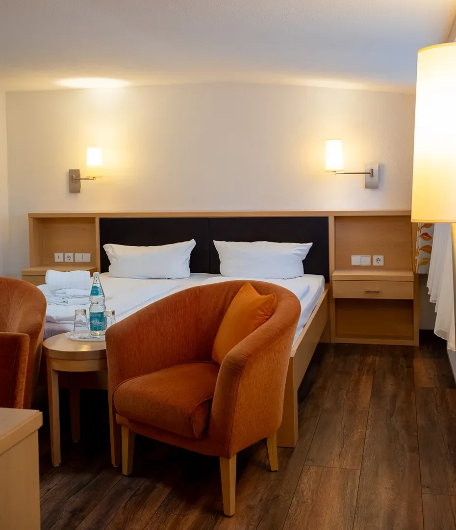 Hotelzimmer mit Doppelbett, zwei orangen Sesseln, Nachttisch und Wandlampen