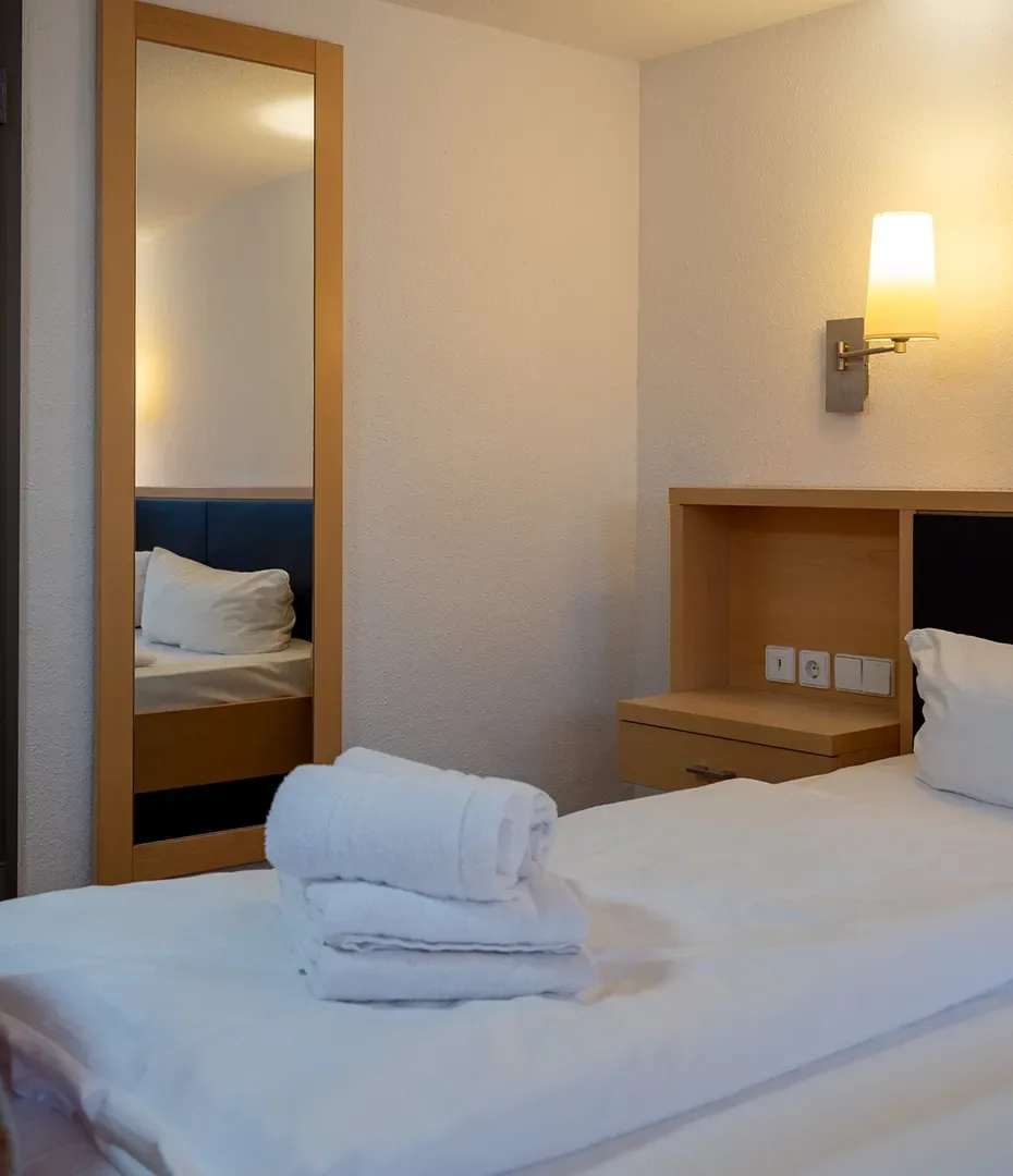 Einfach eingerichtetes Hotelzimmer mit Einzelbett, Wandspiegel, Nachttisch mit Steckdosen und einer Wandlampe, auf dem Bett liegen gefaltete weiße Handtücher