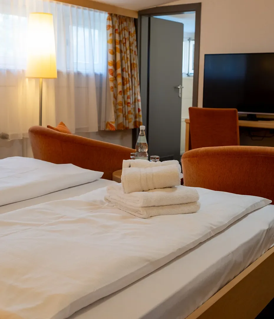 Hotelzimmer mit Bett, auf dem gefaltete Handtücher und eine Wasserflasche stehen, im Hintergrund zwei orangefarbene Sessel, ein Fernseher und ein Fenster mit Vorhängen