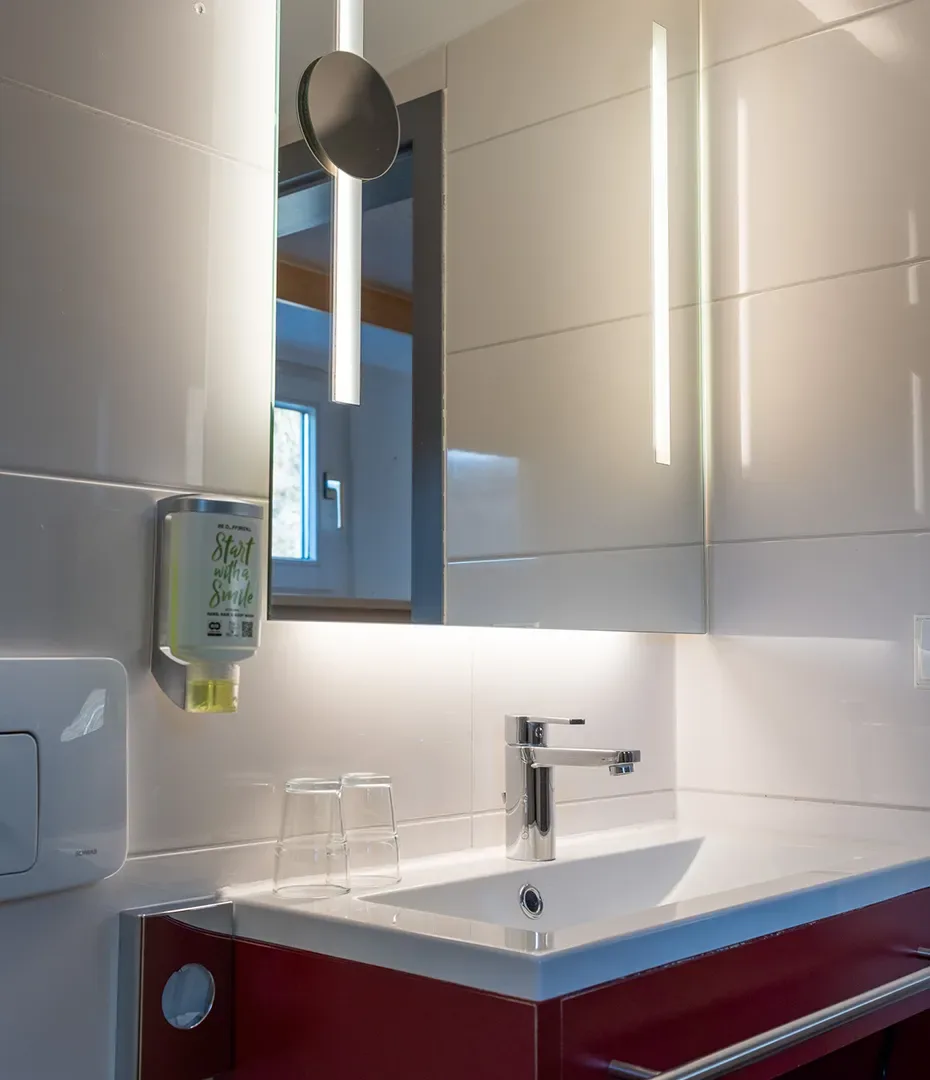 Waschbecken mit Einhebelmischer, zwei Gläsern, Spiegelschrank mit integrierter Beleuchtung und Seifenspender an der Wand in einem Badezimmer unter einer Dachschräge