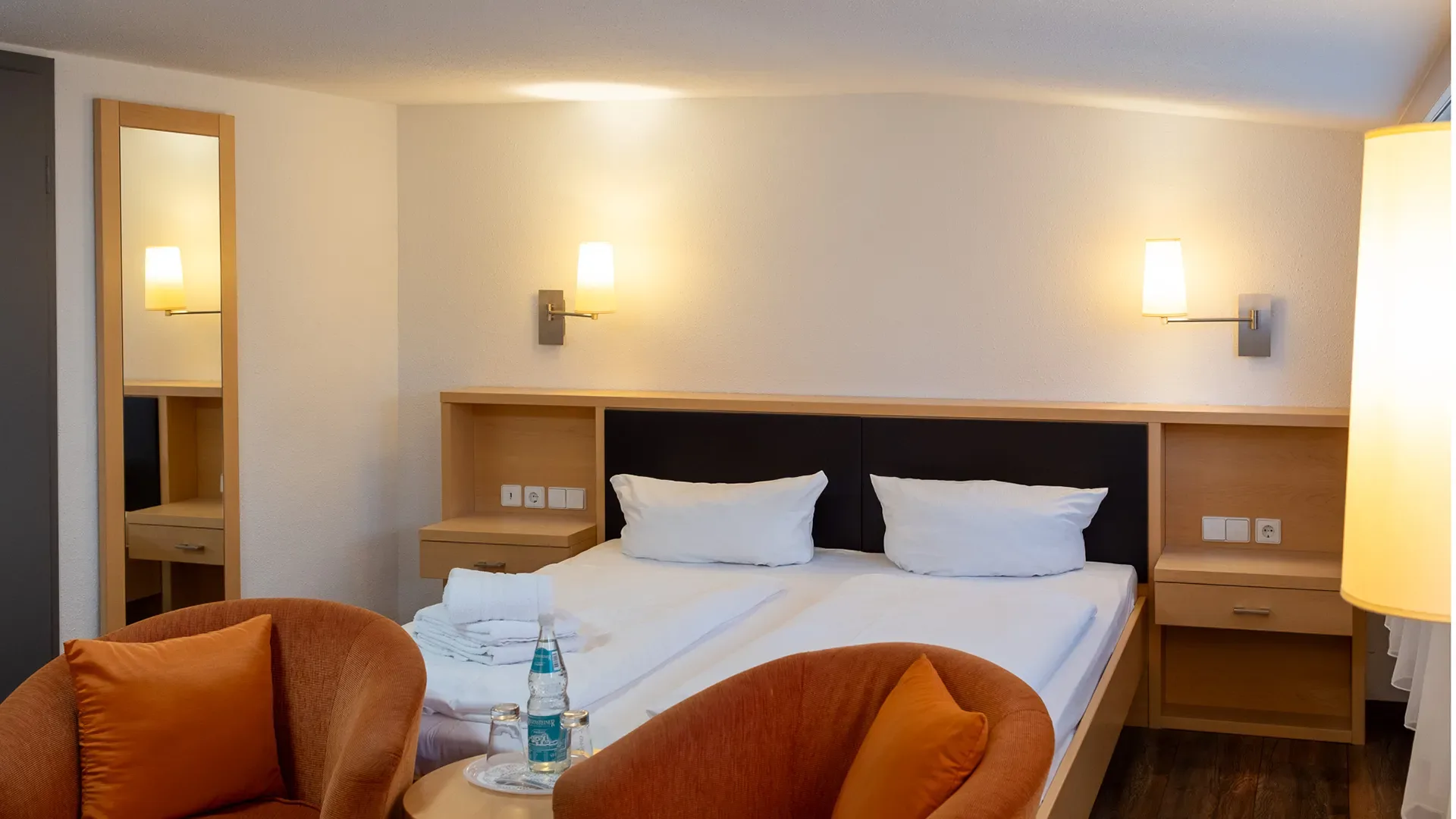 Doppelbett mit weißen Kissen und Bettwäsche in einem Zimmer mit Dachschräge, zwei braunen Sesseln und einem großen Stehlampenlicht