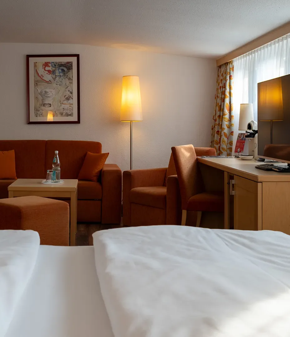 Hotelzimmer mit weißem Bett, orangefarbenen Sesseln, Stehlampe, Schreibtisch mit Fernseher und Fenster mit Vorhängen