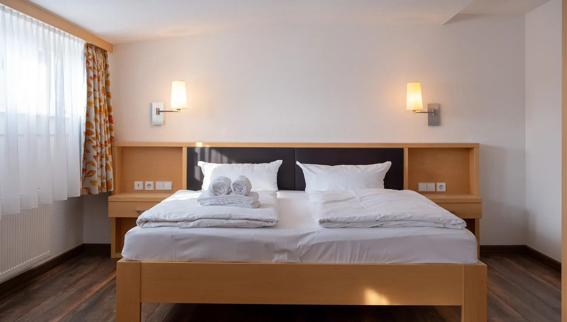 Doppelbett mit weißen Kissen und Bettwäsche in einem hellen Zimmer mit Holzboden, zwei Wandlampen und Fenster mit gemusterten Vorhängen