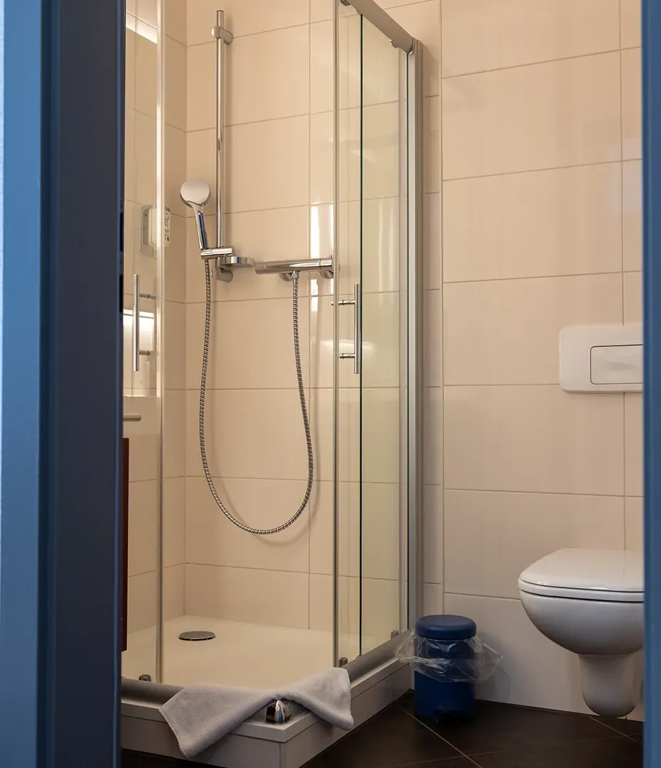 Dusche mit Glasabtrennung, Handbrause und weißem Handtuch auf der Duschwanne neben einer Wandtoilette und einem blauen Mülleimer