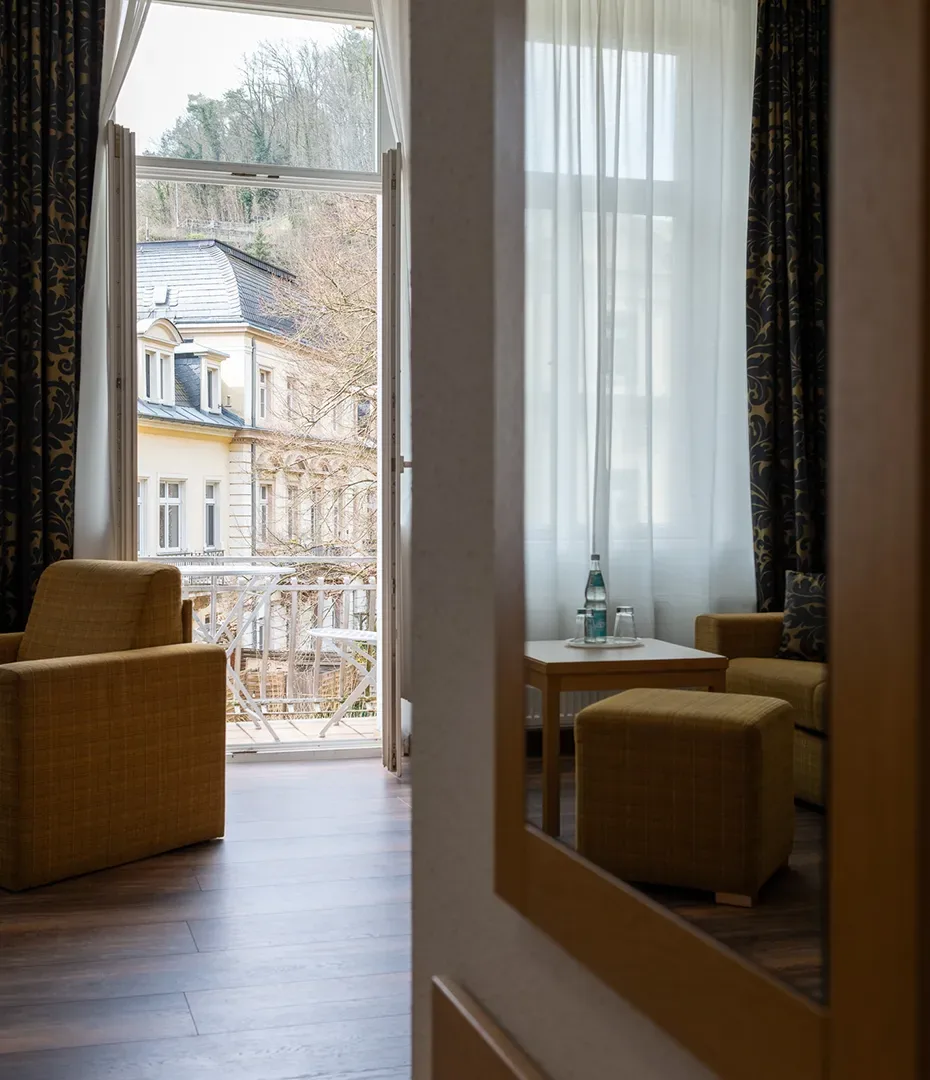 Blick durch einen Spiegel auf einen Balkon mit zwei Sesseln, einem Hocker und einem kleinen Tisch mit Wasserflasche vor einem Fenster mit weißen Vorhängen