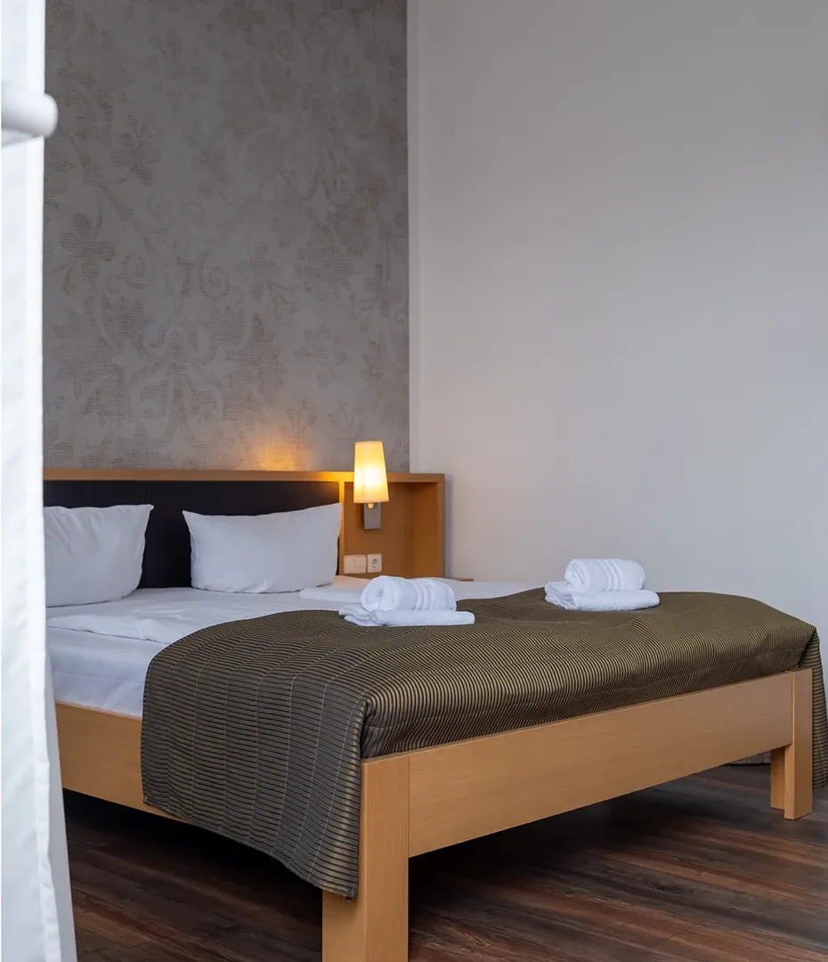 Doppelbett mit zwei weißen Kissen, braunem Bettüberwurf und zwei aufgerollten weißen Handtüchern in einem schlichten Hotelzimmer
