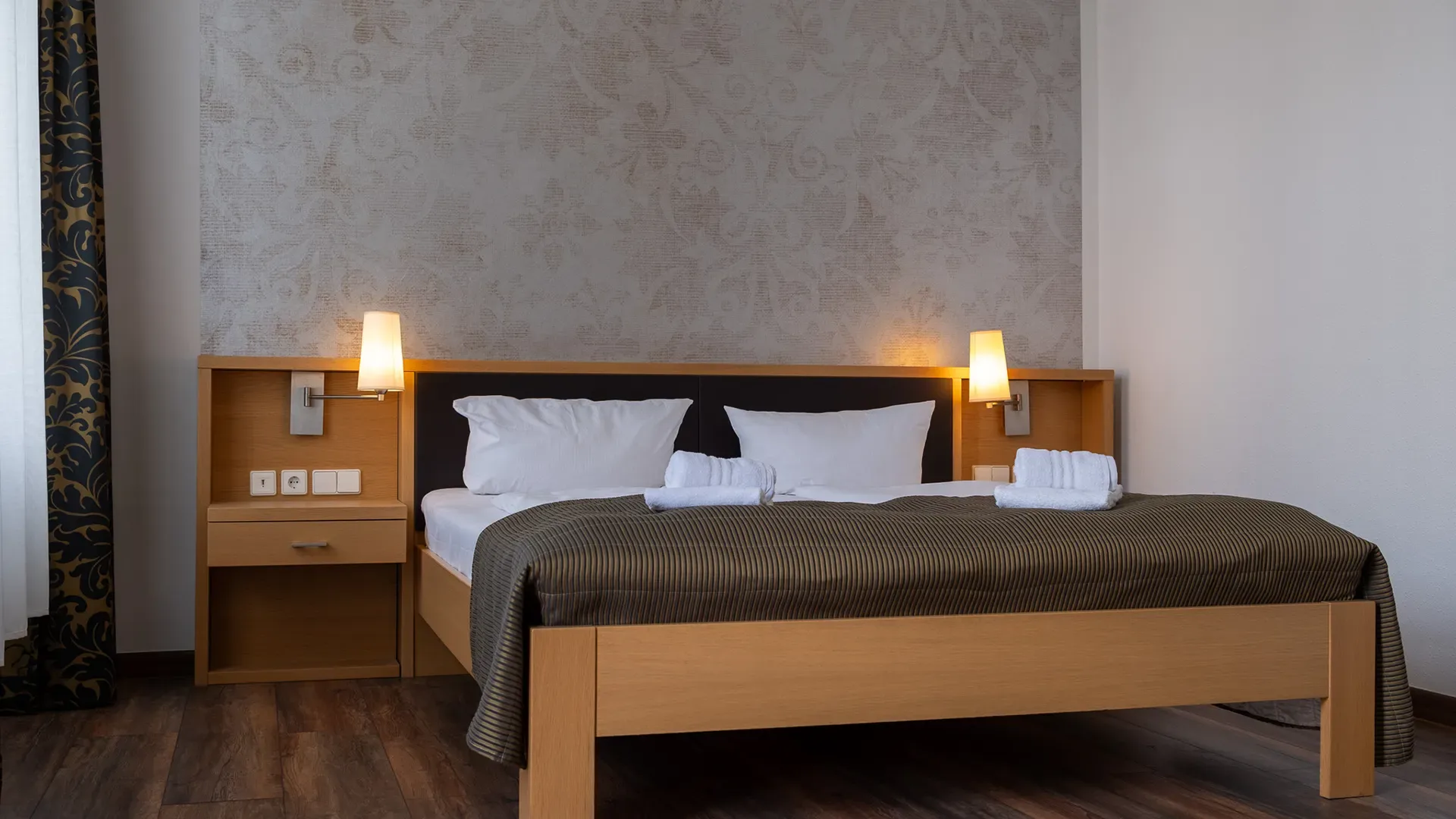 Doppelbett mit weißen Kissen und Handtüchern auf braunem Bettüberwurf in schlicht eingerichteter Hotelzimmerumgebung mit Nachttischen und Wandlampen