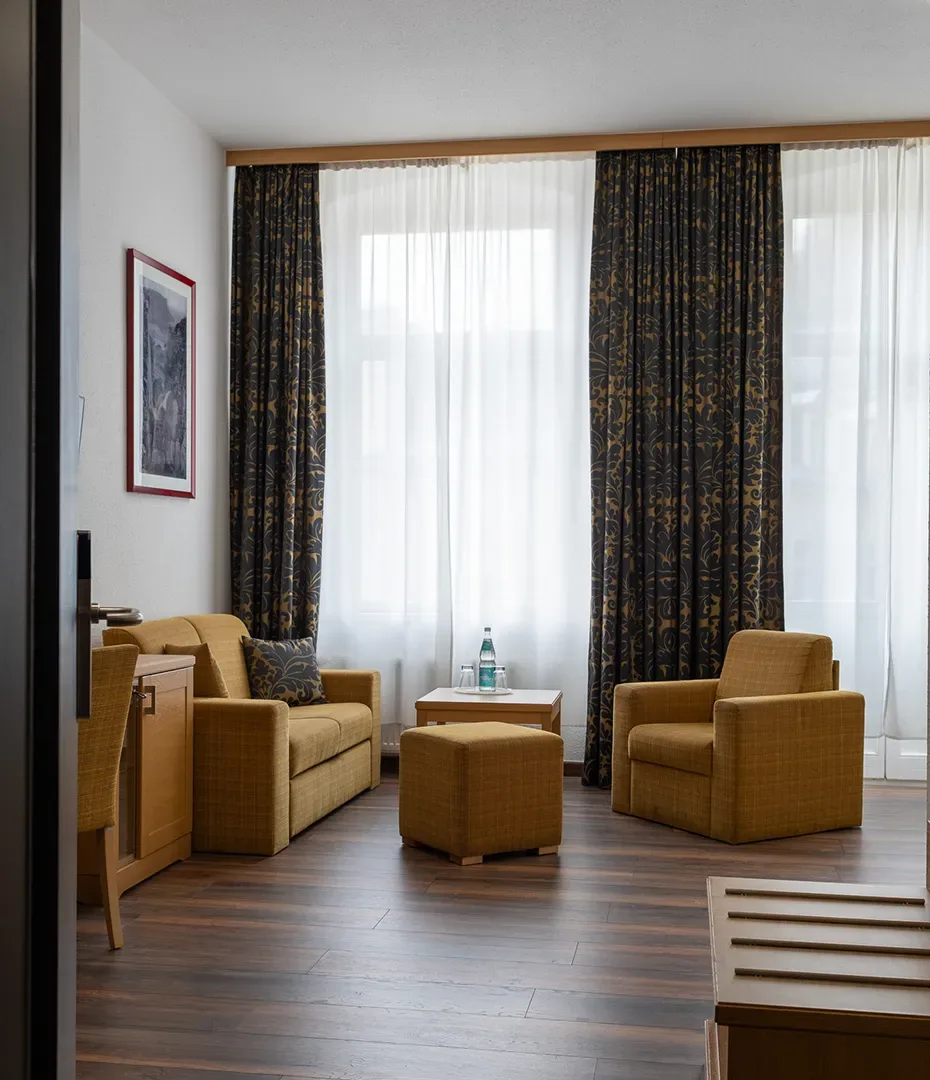 Wohnbereich mit zwei Sesseln, Sofa, Hocker, kleinem Tisch mit Wasserflasche und großen Fenstern mit gemusterten Vorhängen