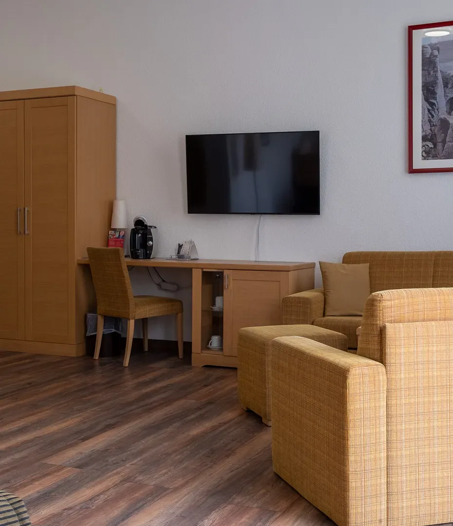 Wohnbereich mit braunem Sofa, Schreibtisch mit Stuhl, Fernseher an der Wand und kleinem Schrank