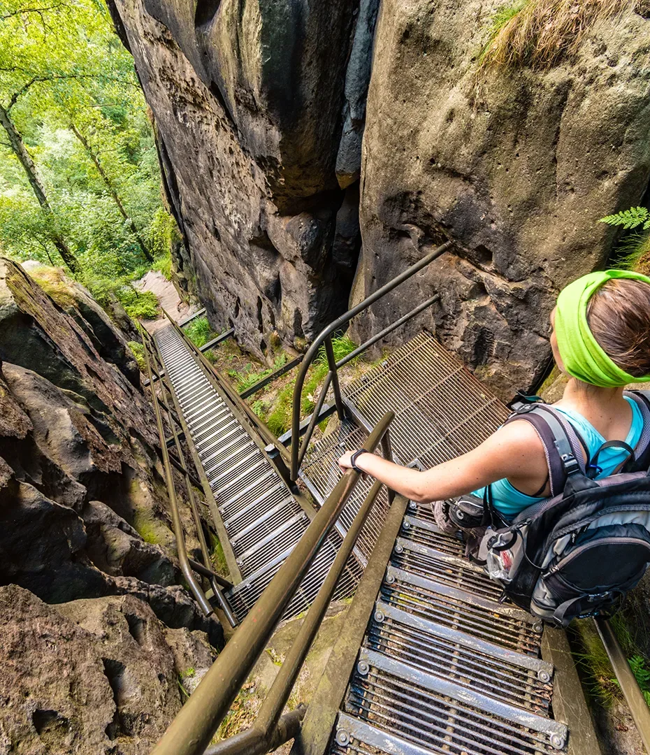 Frau mit grünem Stirnband und Rucksack steigt eine steile Metalltreppe zwischen Felsen im Wald hinab