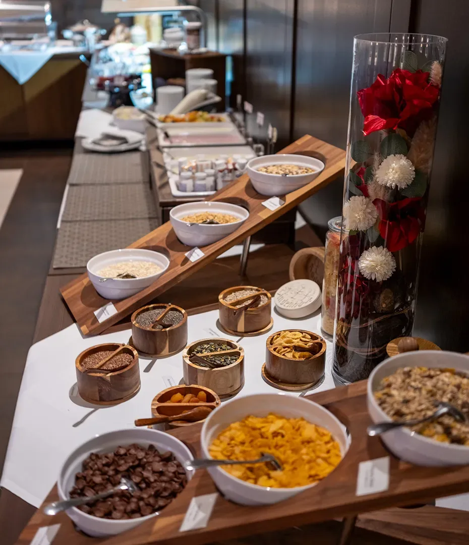 Buffet mit verschiedenen Müslis und Trockenfrüchten auf Holzregalen neben einer hohen Glasvase mit roten und weißen Blumen