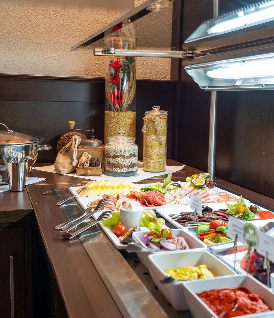 Buffet mit verschiedenen Frühstückszutaten wie Aufschnitt, Gemüse, Saucen und Getreide in Gläsern auf einem dunklen Holztisch