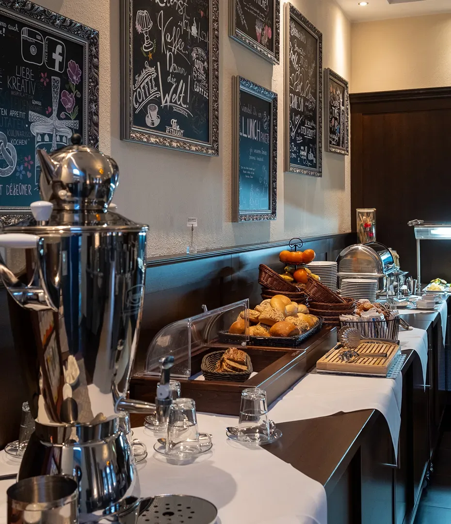 Buffet mit Kaffeeautomat, Gläsern, Brötchen, Obst und weiteren Frühstücksangeboten in einem Raum mit dekorativen Tafeln an der Wand